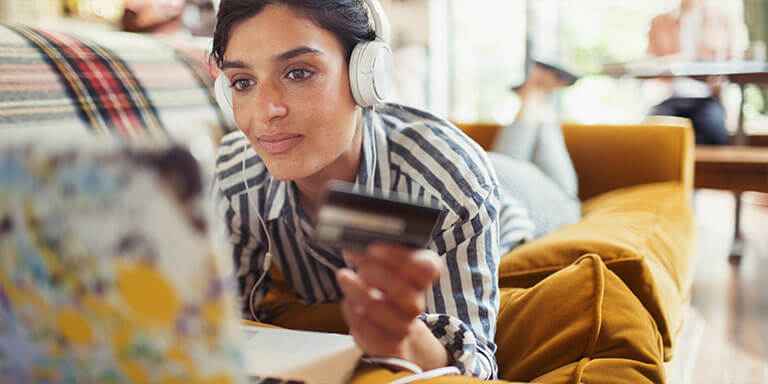 A young woman on her couch with headphones on, staring at her computer while holding a credit card in her hand