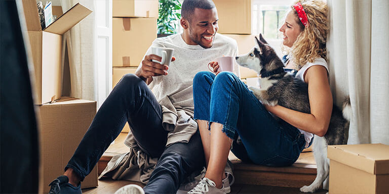 A couple drinking coffee with their husky puppy in a house full of moving boxes