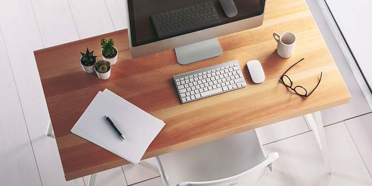 Overhead of a wooden desk with a monitor, keyboard, mouse, papers, coffee, and a pair of glasses on it