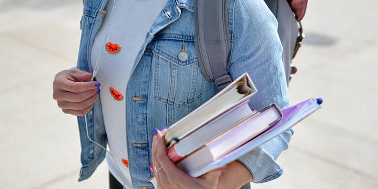 A young women in a jean jacket wearing headphones and carrying her school books 