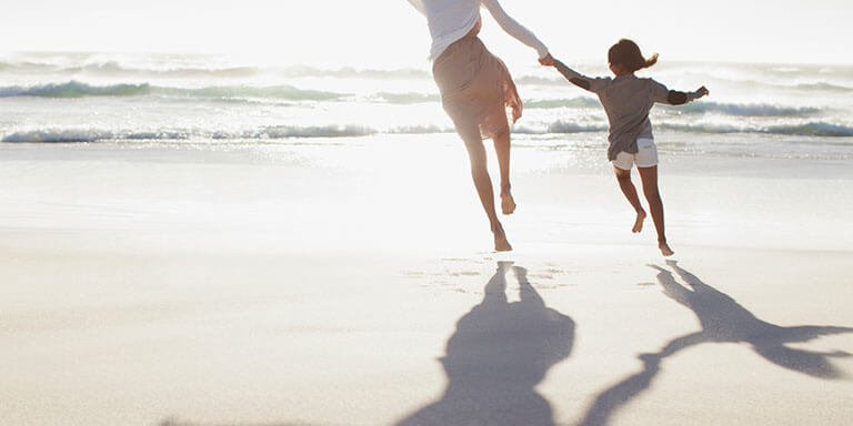 A mother and her young daughter holding hands and running on the beach at sunset