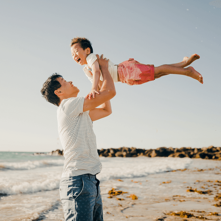 A father laughing and swinging his young son into the air on the beach 