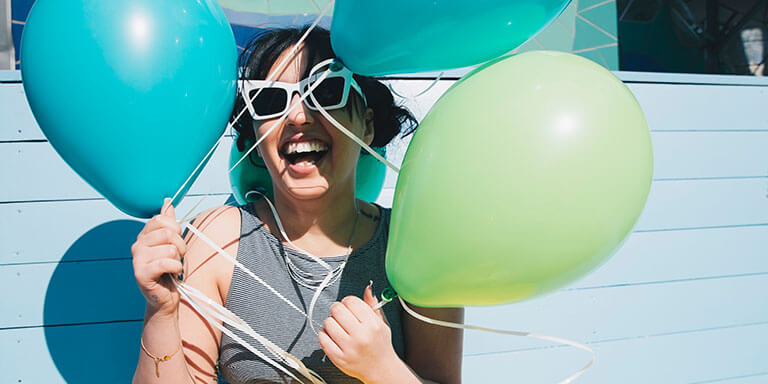 A woman in white sunglasses laughing against a blue house holding a bundle of balloons 