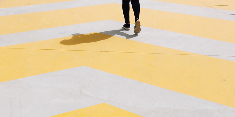 A woman wearing black athletic sneakers walking on a yellow striped concrete sidewalk