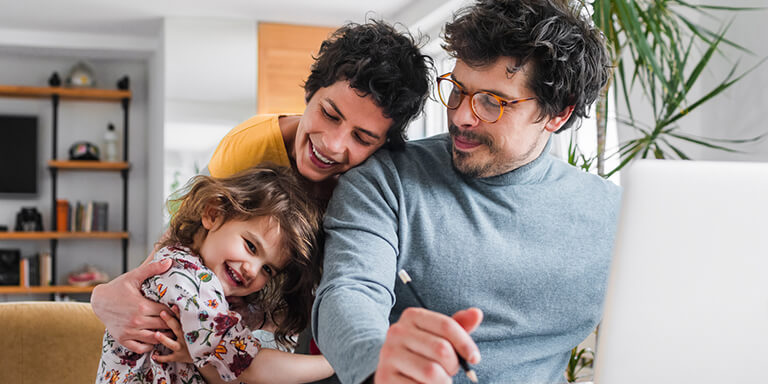 A daughter hugging her mom and dad while her father works at his laptop computer