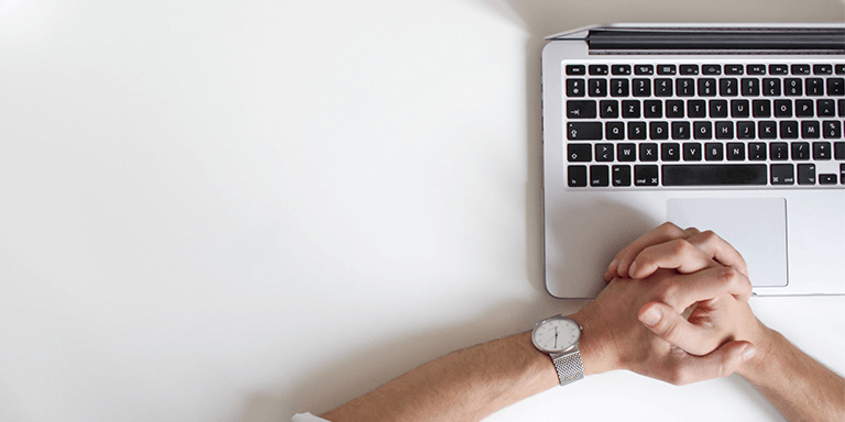 Overhead view of a man wearing a silver watch with his hands clasped over his open laptop