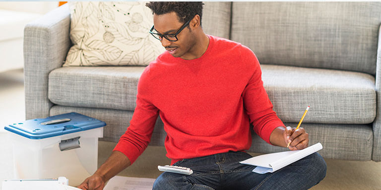 A man in a red shirt working on his computer leaning against his gray couch