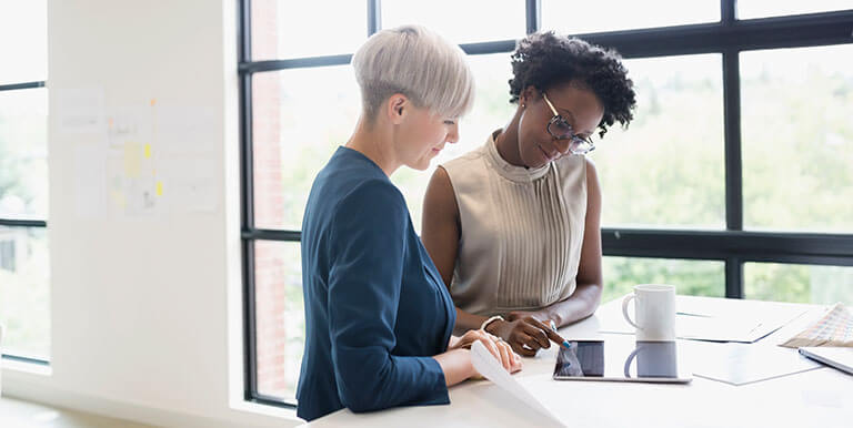 Two women working together on a tablet in an office space at a white table