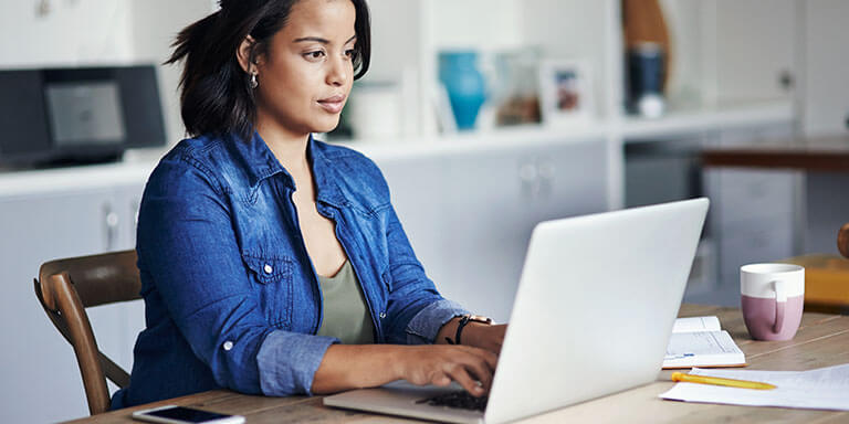 A woman wearing a denim shirt typing on her laptop computer at a wooden table