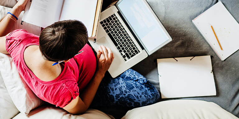 Woman sitting on her couch with her laptop computer, textbooks, and multiple notebooks