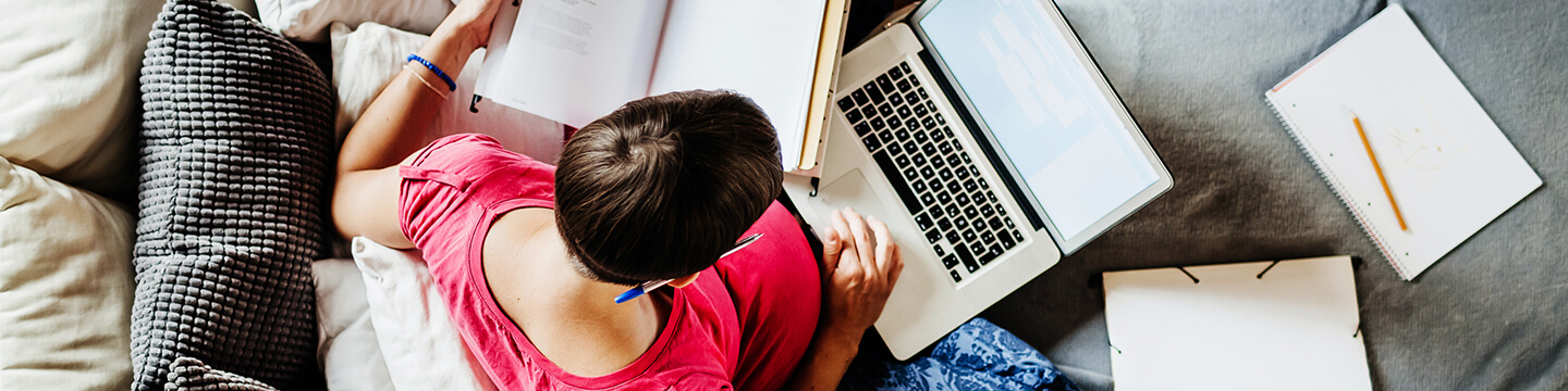 Woman sitting on her couch with her laptop computer, textbooks, and multiple notebooks