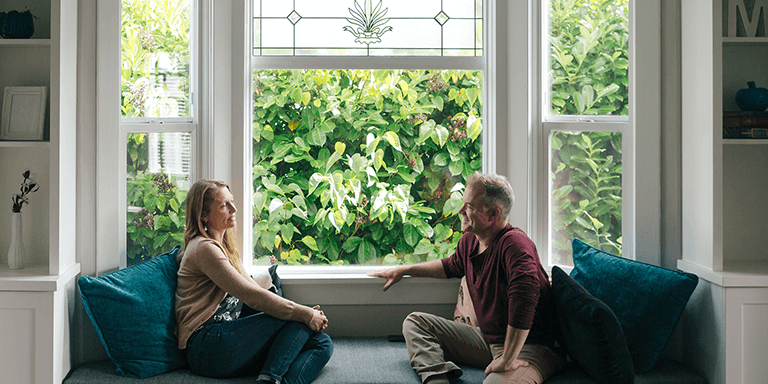A man and woman sitting in their house on a window seat in front of a wall of ivy