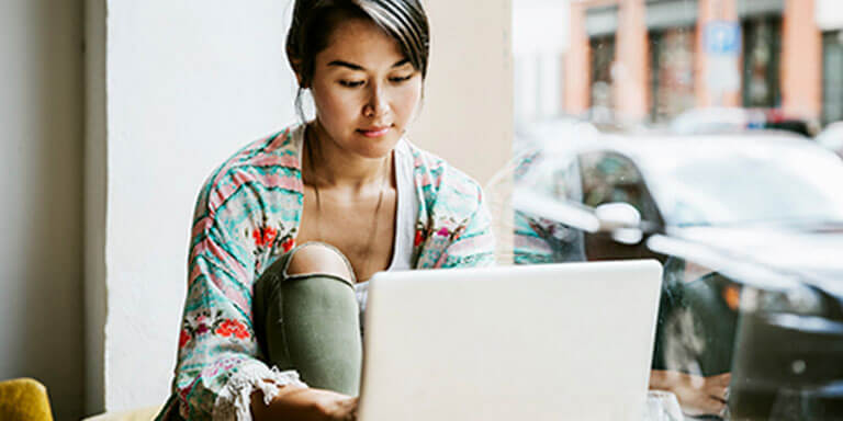 A woman sitting at a table by a window typing on her laptop computer 