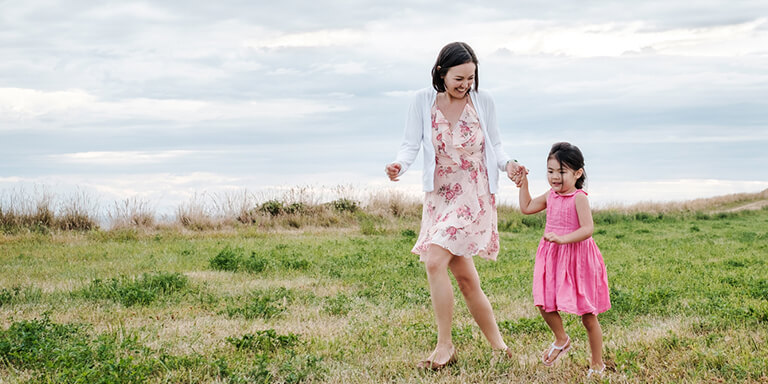A woman and her young daughter in pink dresses running together in thick grass