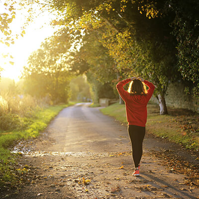 A young woman in athletic gear walking down a tree-lined drive with her hands resting above her head 