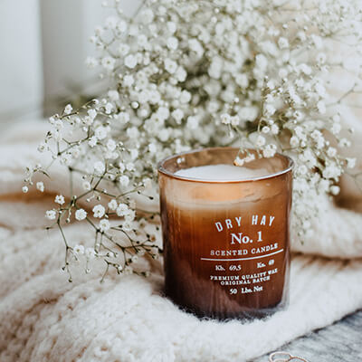 An unlit scented candle resting on a knit white blanket and bouquet of baby's breath