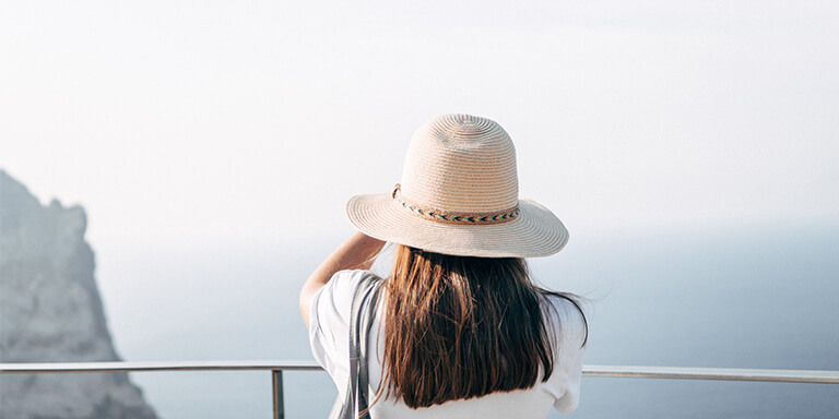 A woman in a sun hat talking a photo of the ocean and cliffside views