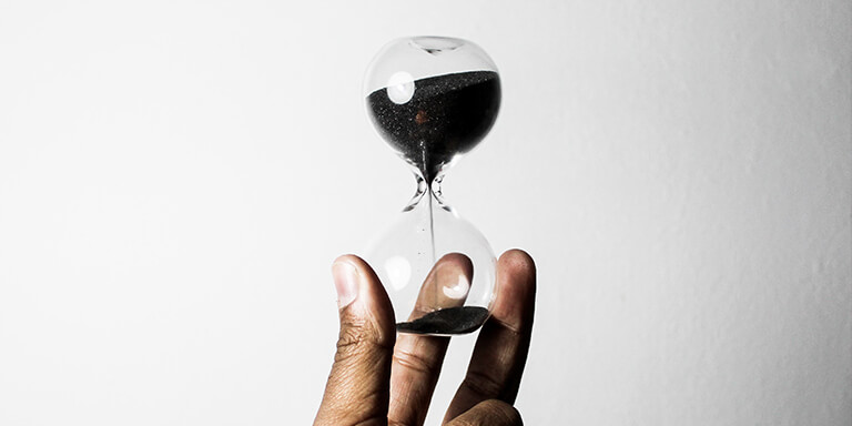 A hand holding up an hourglass with black sand against a white wall 