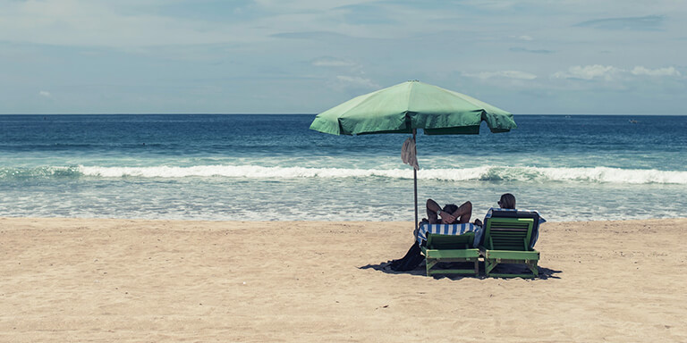 A couple sitting in beach chairs underneath an umbrella facing the ocean on an empty beach 