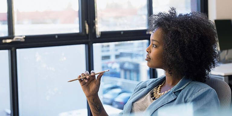 A businesswoman at her desk holding a pencil in her hand and staring out of the window of her office building