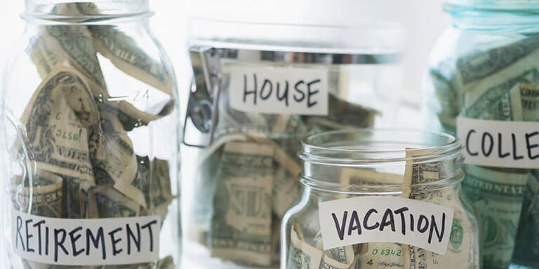 4 Glass jars labeled with "Retirement", and "College", "Vacation",  and filled with dollar bills. 