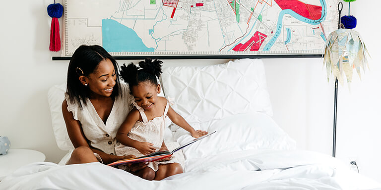 A mother and her young daughter sitting together on her bed and smiling 