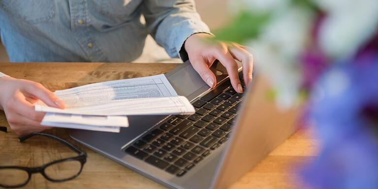 Woman holding tax documents in one hand and typing on her laptop computer keyboard with the other