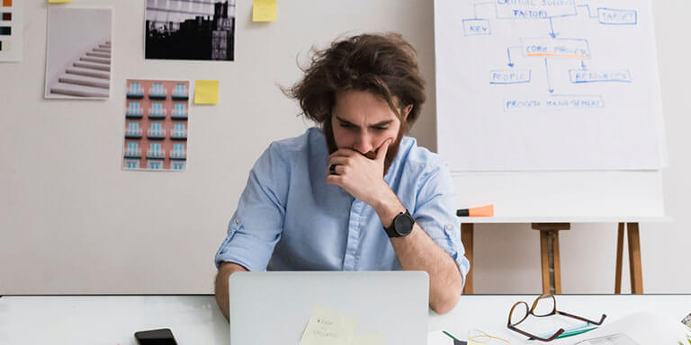 A young man in a blue button up shirt studying something on his laptop with charts on the wall behind him