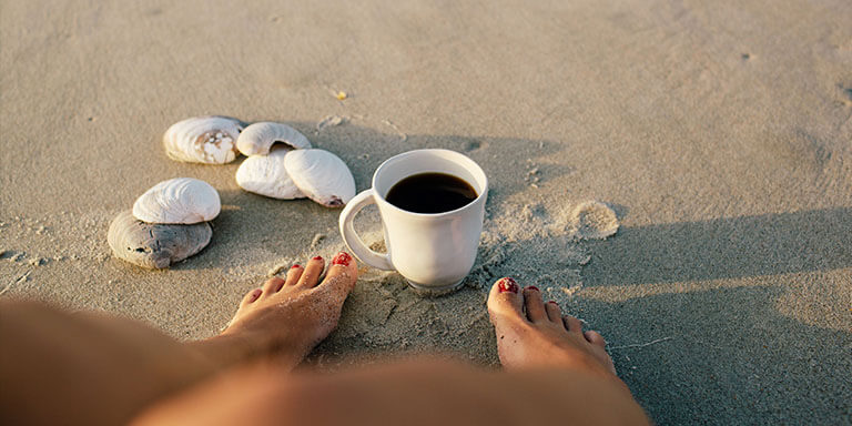 A woman sitting on the beach with a cup of coffee and seashells at her feet