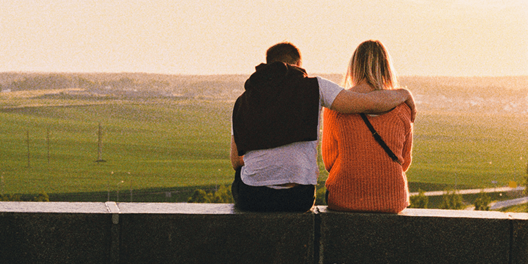 Man with his arm around a woman sitting on a ledge overlooking green fields at sunset 