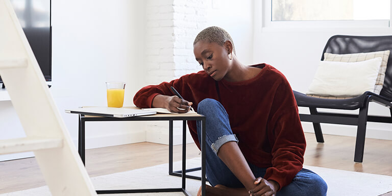 A woman sitting on the floor using a side table as a writing surface with a glass of orange juice next to her