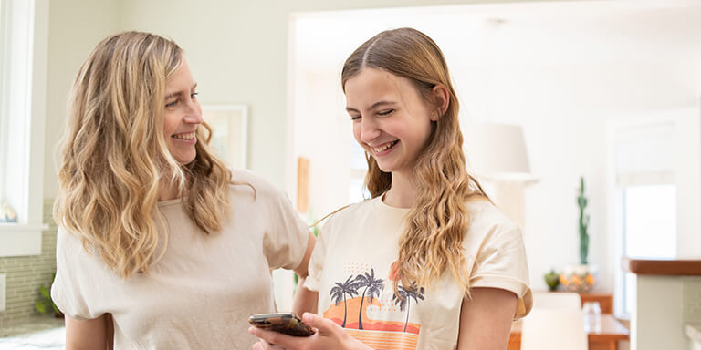 A mom and daughter laughing together at something on the daughter's smartphone