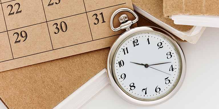 Close up of a silver pocket-watch resting against a cork board calendar