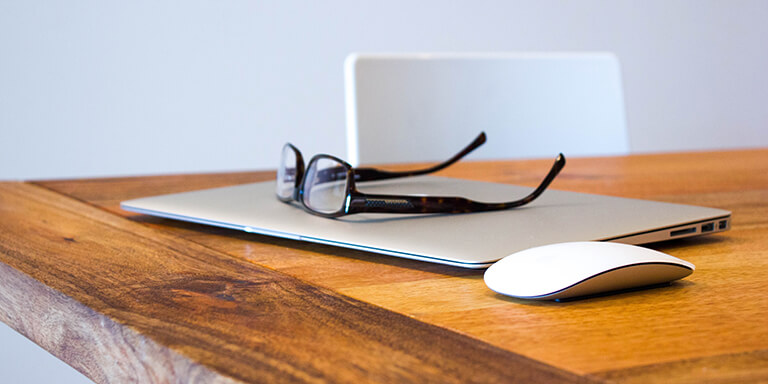 Black reading glasses resting on top of a laptop on a wooden desk next to a mouse