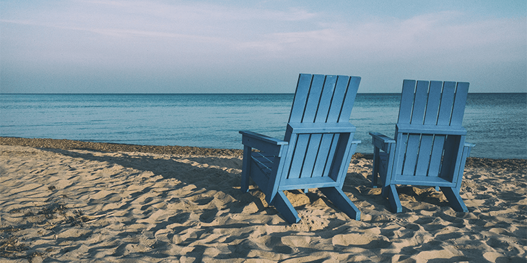 Two blue Adirondack chairs on the beach facing towards the ocean