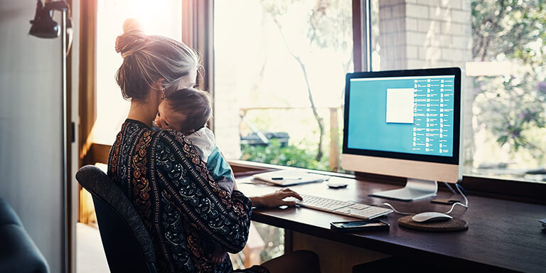 A mother who has her infant child resting on her shoulder while she works on her home computer