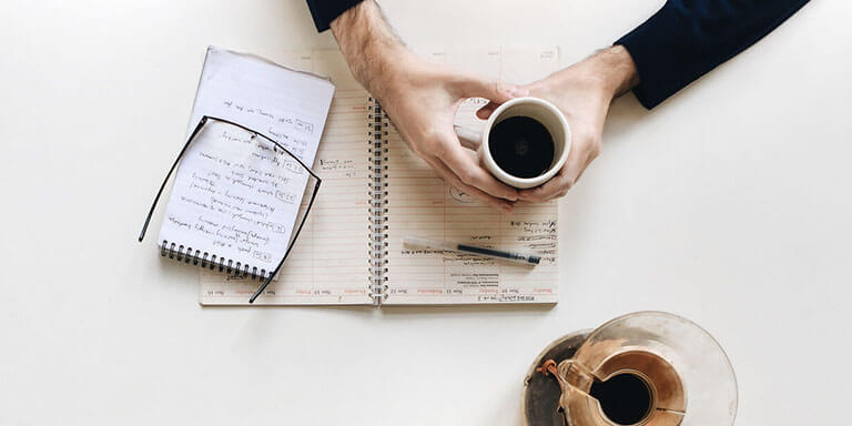 Overhead view of a woman holding a cup of coffee while writing in two journals