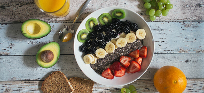 A bowl of assorted cut fruit on a table surrounded by bread, assorted fruits, and a glass of juice