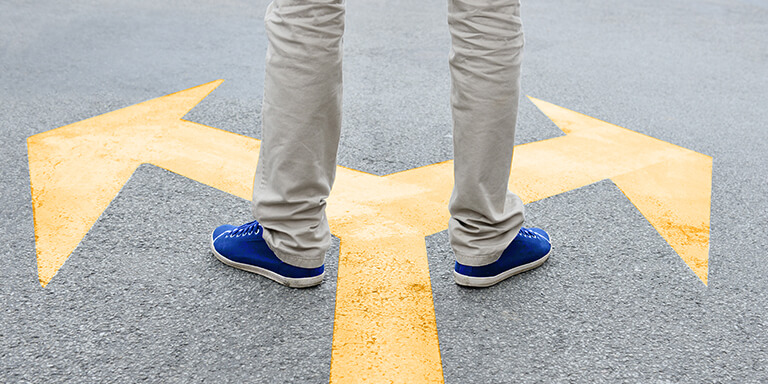 A man standing in front of a sign that indicates an upcoming fork in the road and has one foot on either side