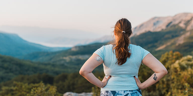 A woman with an insulin pump in her arm standing on the top of a mountain gazing over a valley 
