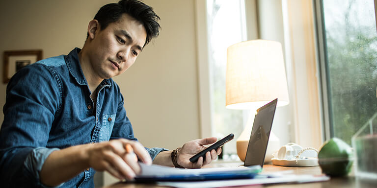 A man sitting at a desk flipping through a notebook while holding his smartphone and looking at something on his laptop computer