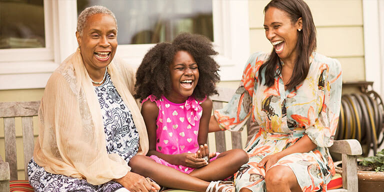 A grandmother, mother, and daughter laughing together on a bench outside of their house