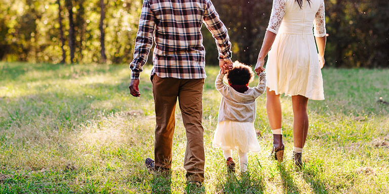 A mother and father holding their young daughters hands in between them as they walk together through a grassy field