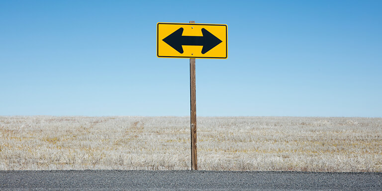 A yellow two direction arrow road sign in an intersection against a blue sign 