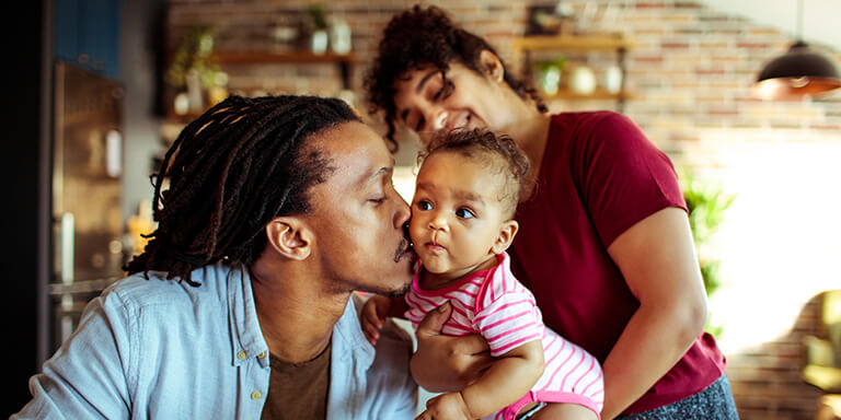 A father kissing his infant daughter on the cheek while her mother looks on and smiles