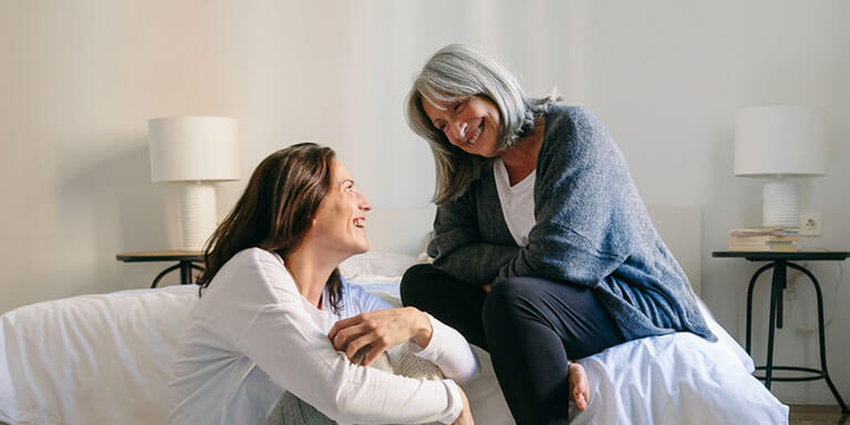 Older woman sitting on her bed while talking to her daughter who is sitting on the floor