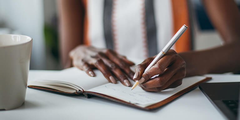 Close up of a woman writing in her open journal with a cup of coffee sitting on the table 