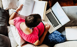 Overhead of a women sitting on a couch, working on her laptop and a notebook at the same time
