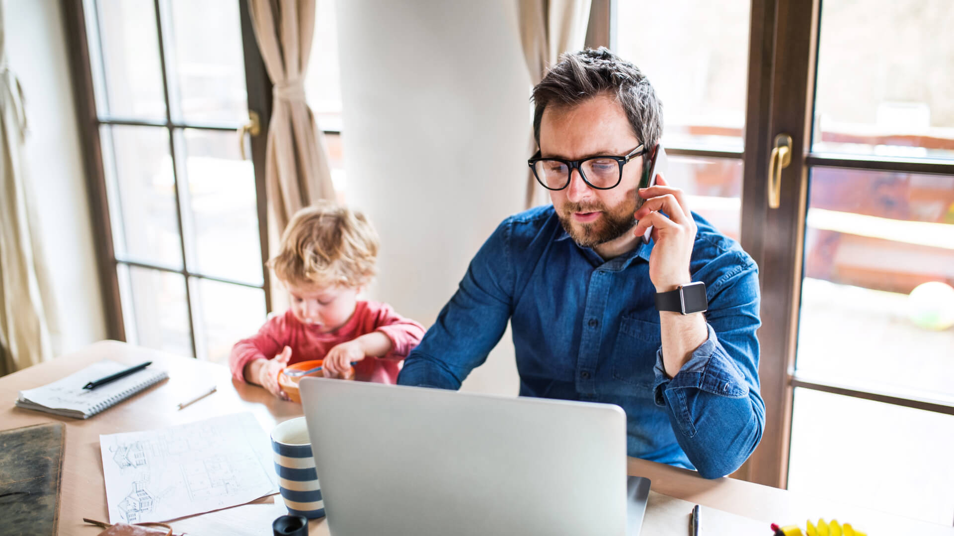 Father on the phone while his young son plays next to him at the table