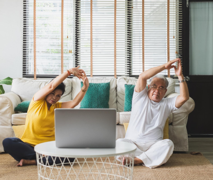 A couple doing yoga on the floor of their living room and watching an online class on their laptop computer.